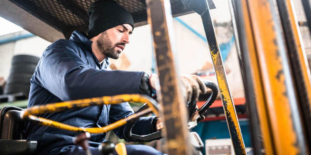A man in a beanie and gloves operates a forklift in an industrial warehouse, focused on controls, showing good fleet management.