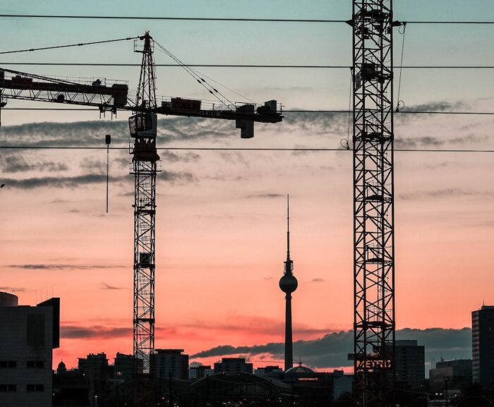 Construction cranes against a pink-blue sunset with city skyline and power lines, suggesting a connected jobsite.