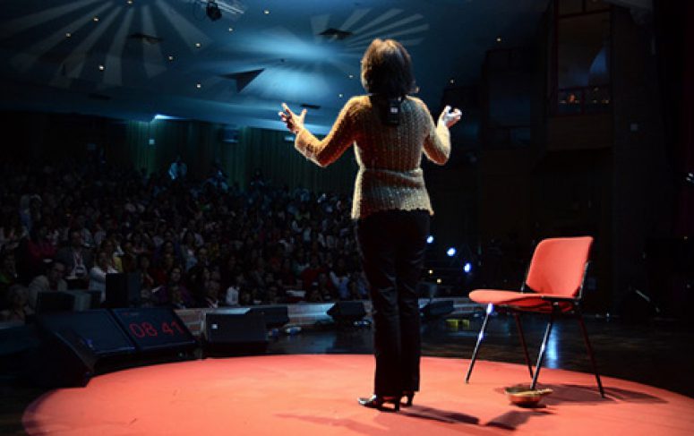 A person gestures on a red stage under bright lights, with a chair and basket nearby—capturing a connected jobsite presentation.