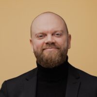 A smiling bearded man in a black turtleneck and blazer stands before a beige background, suggesting construction IoT expertise.