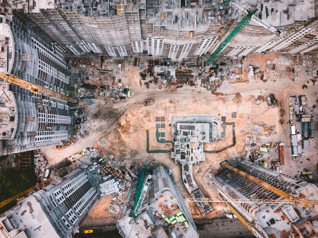 Aerial view of a large construction site with cranes, unfinished buildings, scattered materials, and dirt roads showing a connected jobsite.