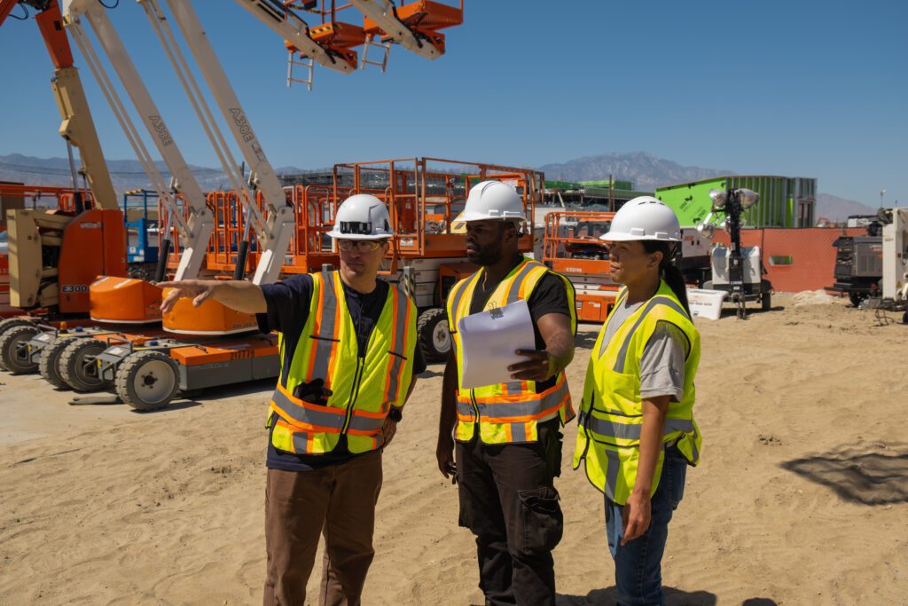 Three construction workers in safety vests and helmets review plans on a sandy site with equipment in the background, showing connected jobsite.