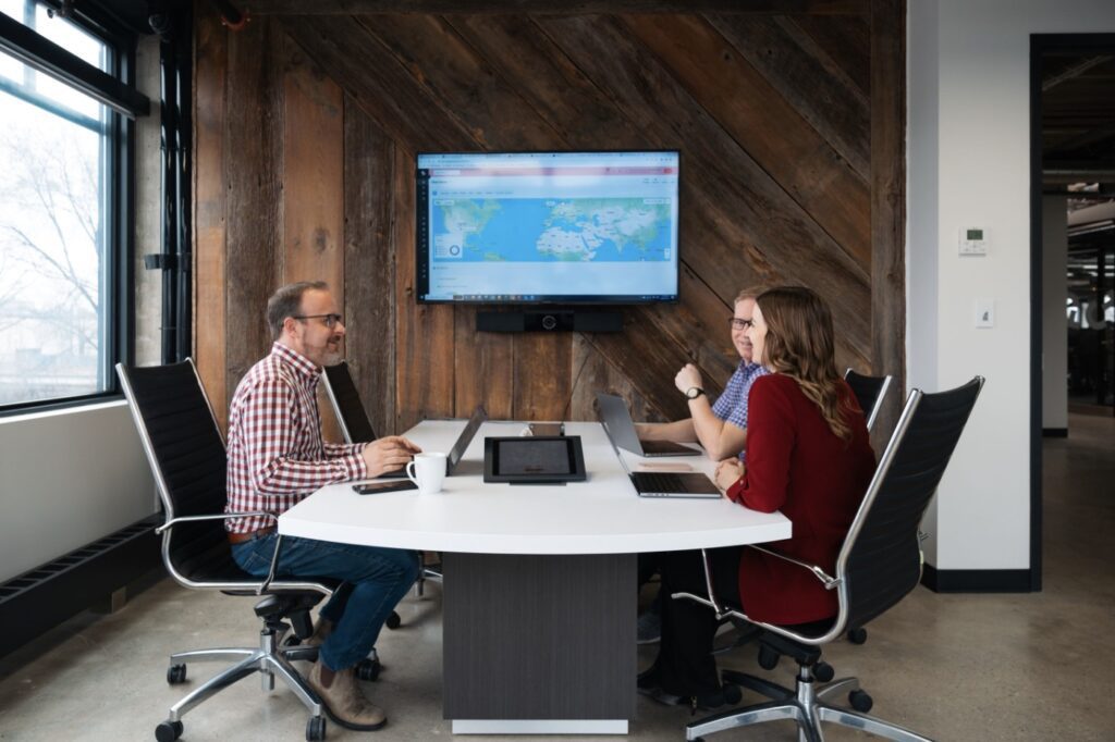 Three people with laptops meet at a modern office table, viewing a world map display—ideal for fleet management discussions.