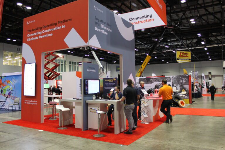 A red and white trade show booth with Trackunit displays, digital screens, and people talking about construction equipment tracking.