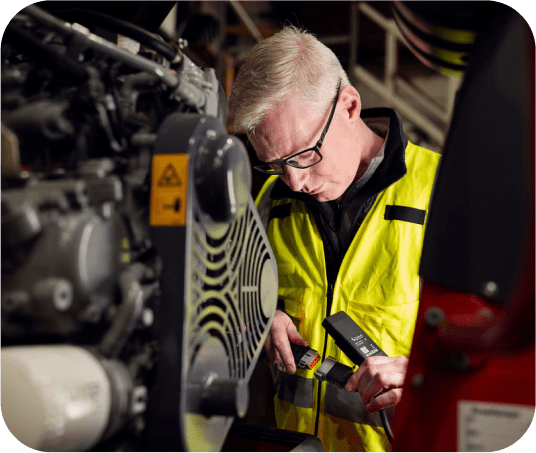 A man in a yellow safety jacket inspects machinery with a diagnostic tool in an industrial setting, showing construction IoT in action.