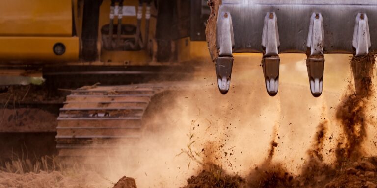 Close-up of an excavator bucket digging into dry soil, dust flying, with yellow body and tracks behind; ideal for construction IoT.