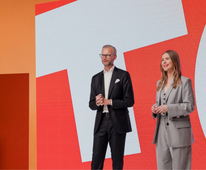 A man in a black suit and a woman in a gray suit smile on stage before a bold geometric backdrop, at a connected jobsite event.