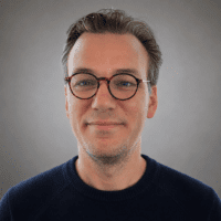 A man with short brown hair and round glasses, wearing a dark blue sweater, smiles at the camera in front of a light gray background.