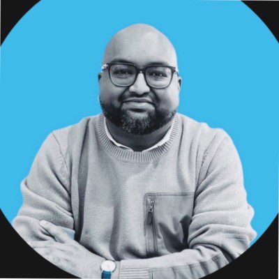 A man with glasses and a beard in a light sweater sits smiling with arms crossed, against a bright blue background; fleet management.