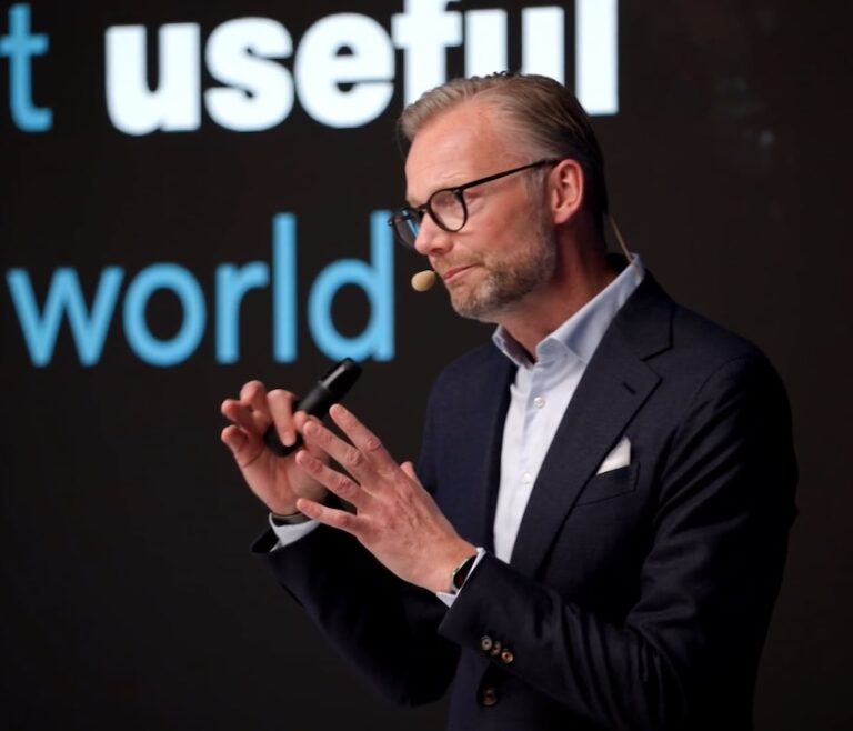 A man in a suit and glasses speaks on stage about safe equipment access, with projected text behind him, related to construction IoT.