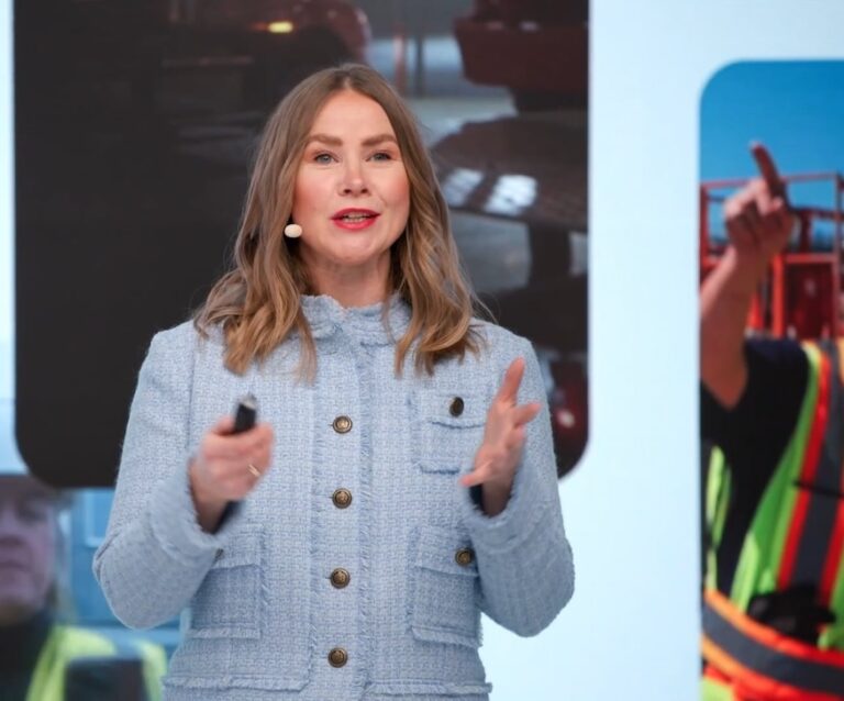 A woman in a light blue jacket speaks on stage about safe equipment access, with safety vest workers behind her, highlighting fleet management.