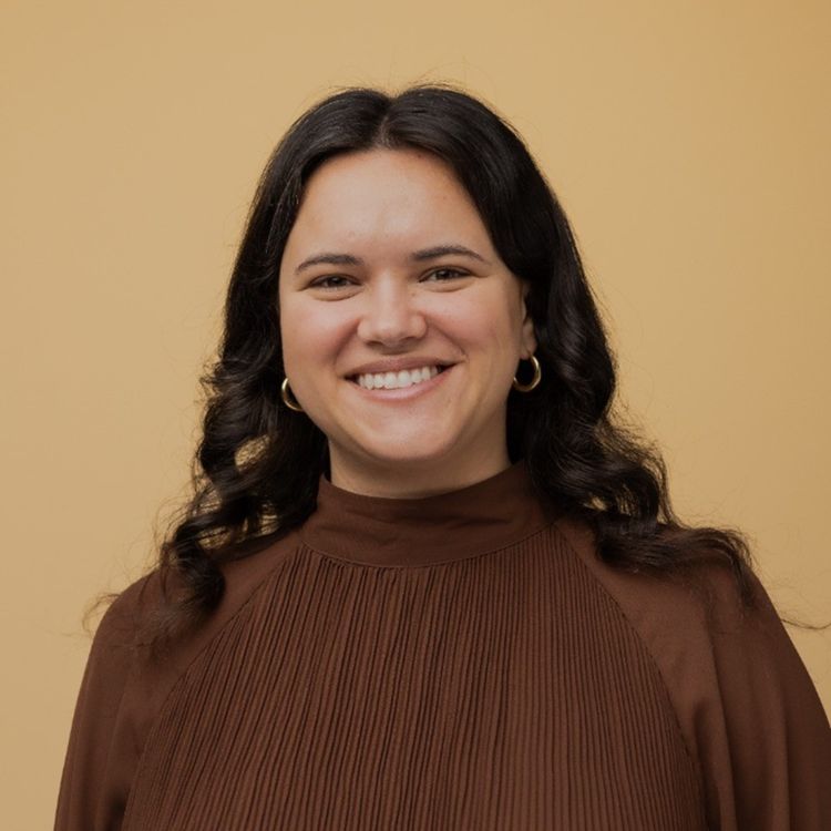 A woman with long dark wavy hair in gold hoop earrings and a brown pleated top smiles before a light tan background, suggesting telematics.