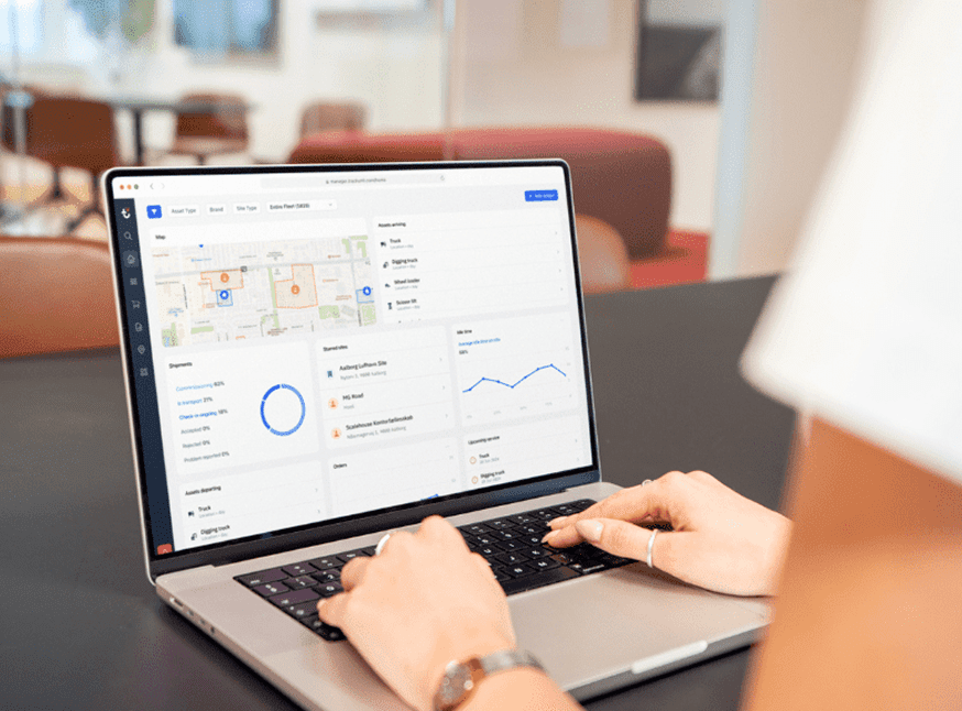 Person using a laptop displaying a dashboard with a map, charts, and data graphs on the screen, seated at a table in a modern office environment.