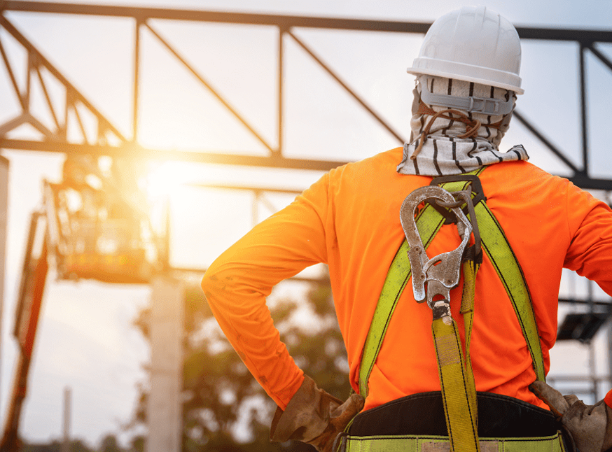 A construction worker wearing a hard hat, safety harness, and bright orange shirt stands with hands on hips, looking at a metal structure at a construction site during sunset.