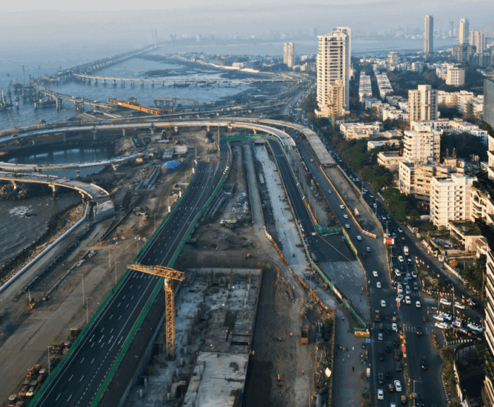 Aerial view of a coastal city with high-rises, busy roads, ongoing construction using Trackunit IrisX, and a curved sea bridge.