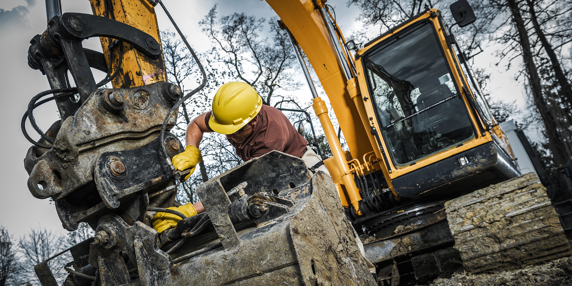 A worker in a yellow hard hat repairs an excavator bucket with aftermarket parts at a muddy site, highlighting construction equipment tracking.