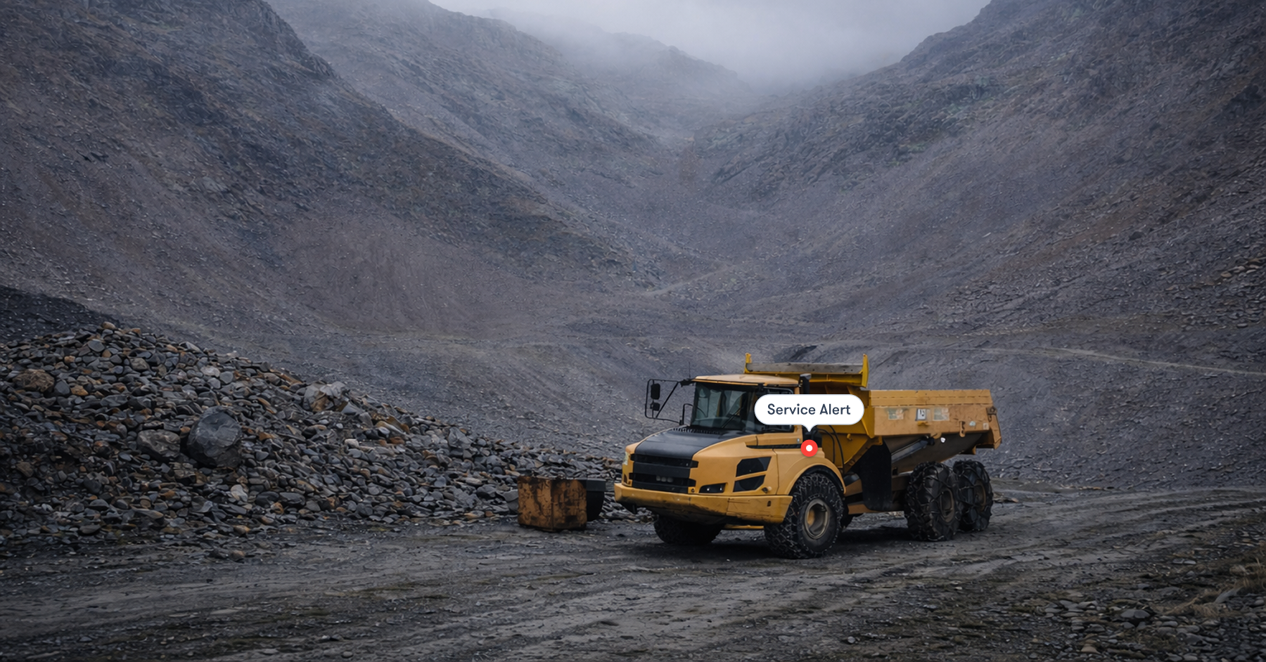 A yellow dump truck with aftermarket parts is parked on a rocky quarry road under a Service Alert, showing construction IoT in action.