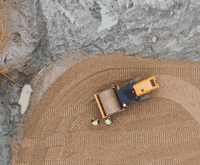 Aerial view of a connected jobsite with a yellow bulldozer moving gravel, two workers in green vests, and tire tracks in the dirt.