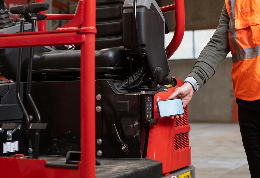 A person in an orange safety vest uses a smartphone near a black and red construction vehicle, suggesting construction IoT use.