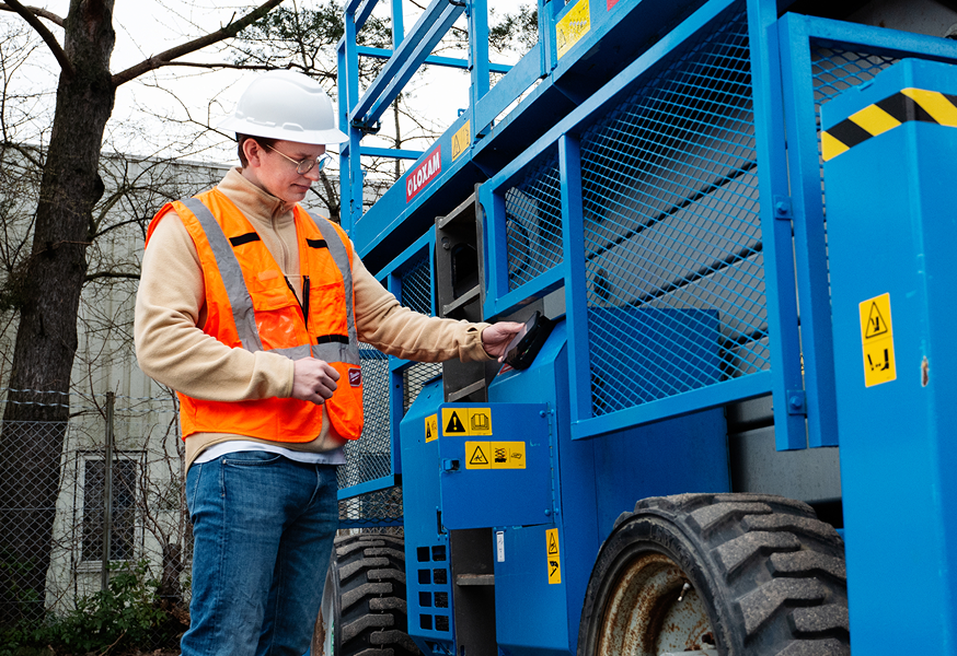 A construction worker in safety gear operates a blue scissor lift outdoors, showcasing connected jobsite technology. Trees in background.