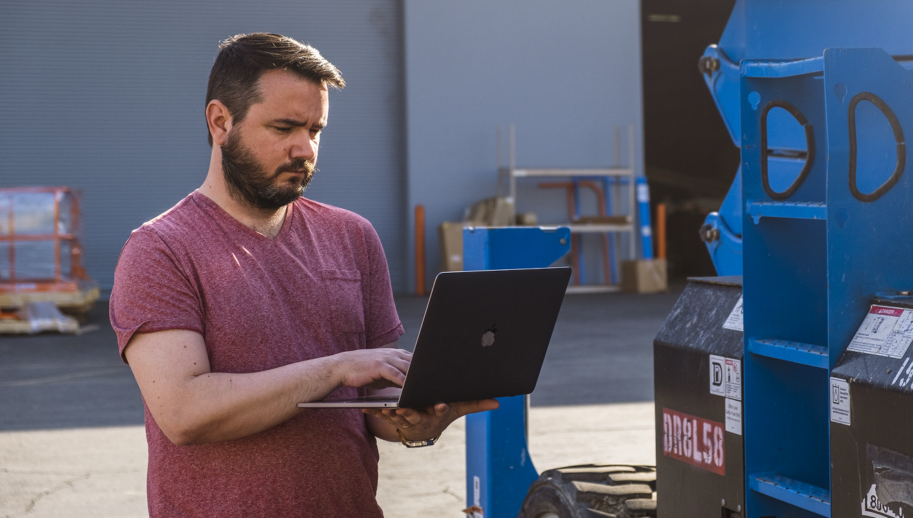 A bearded man in a red t-shirt uses a laptop outdoors by industrial equipment and pallets, suggesting construction equipment tracking.