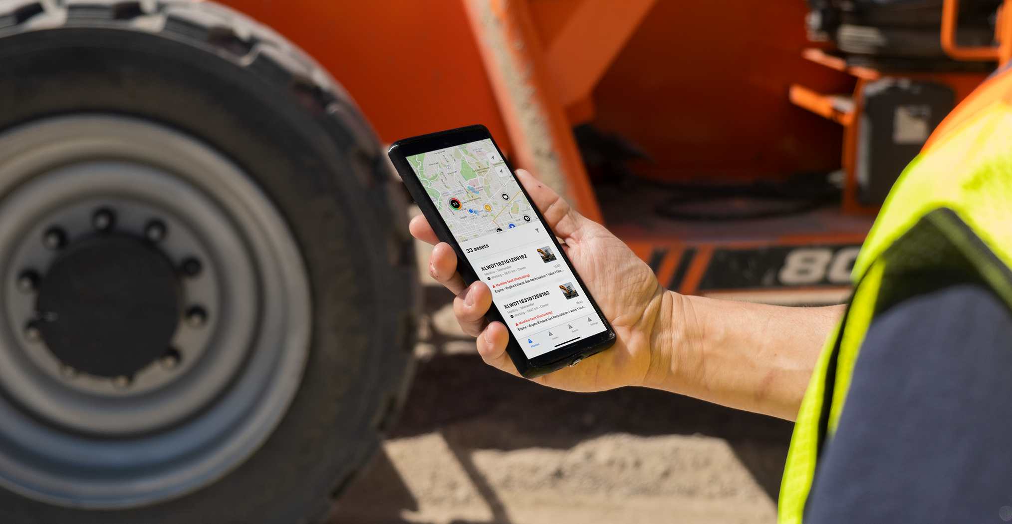 A person in a high-visibility vest holds a smartphone with a map and job info near construction equipment, showing fleet management.
