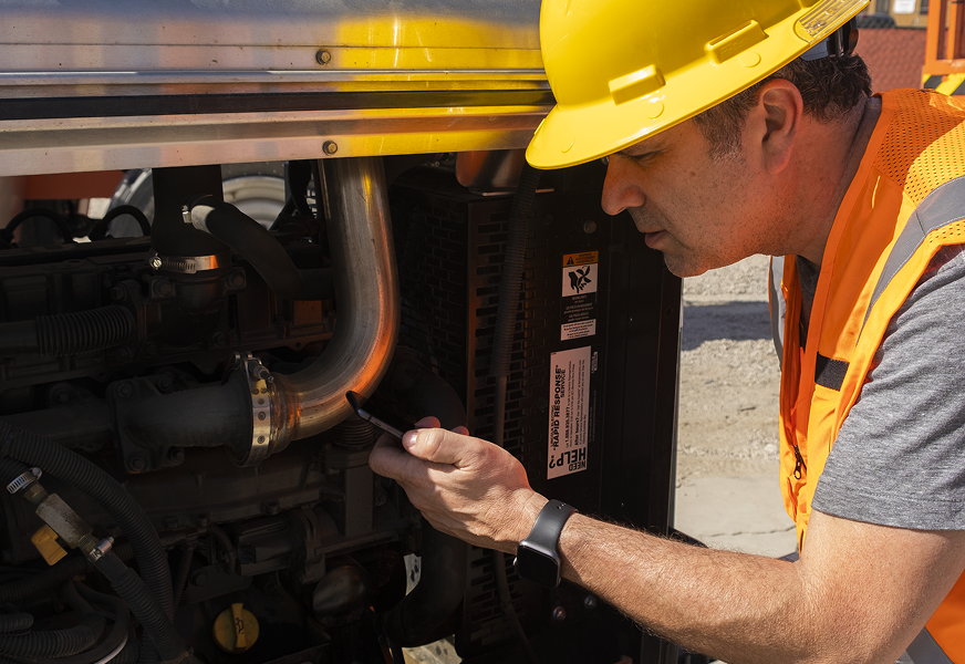 A construction worker in a hard hat and safety vest inspects a vehicle engine outdoors, supporting construction equipment tracking.