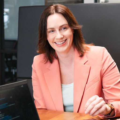 A woman in a peach blazer smiles at a laptop showing Trackunit IrisX, seated at a wooden table in an office for construction IoT.