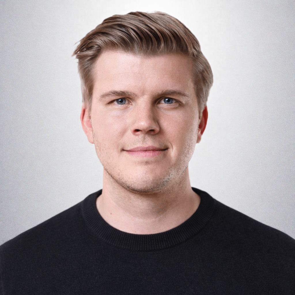 A young man in a black Trackunit IrisX sweater smiles slightly in front of a plain gray background, suggesting construction IoT.