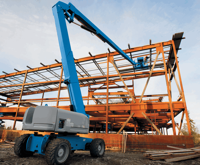 A blue boom lift works by a steel building frame at a connected jobsite, with scattered materials and trees in the background.