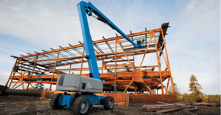 A blue boom lift works by a steel building frame at a connected jobsite, with scattered materials and trees in the background.