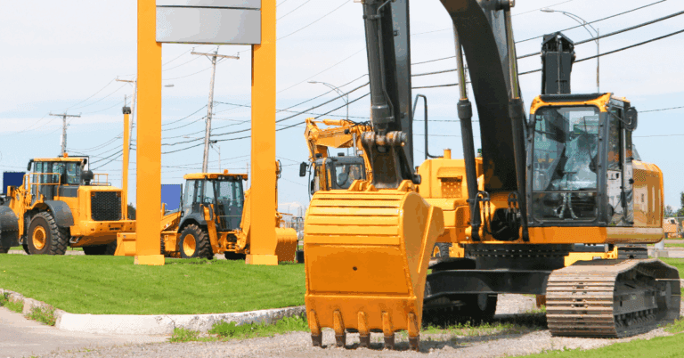 Yellow construction vehicles, including an excavator and loaders, are parked on gravel near a street—fleet management.