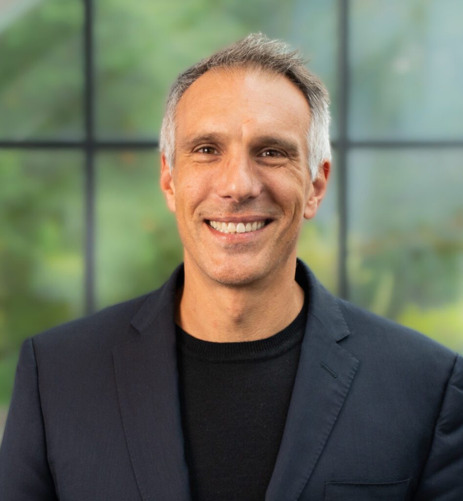 A man with short gray hair in a dark blazer smiles at the camera, window and greenery behind him, suggesting a fleet management setting.
