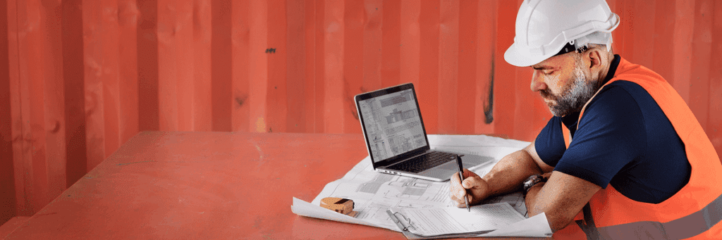 A construction worker in a hard hat and safety vest reviews blueprints at a desk with a laptop, showcasing a connected jobsite.