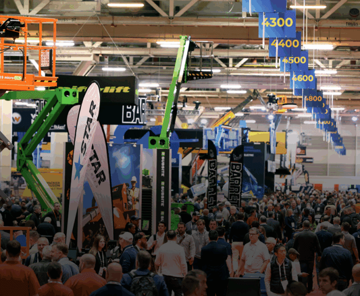 A crowd walks through bauma 2025's indoor trade show, surrounded by construction equipment and banners, highlighting connected jobsite.