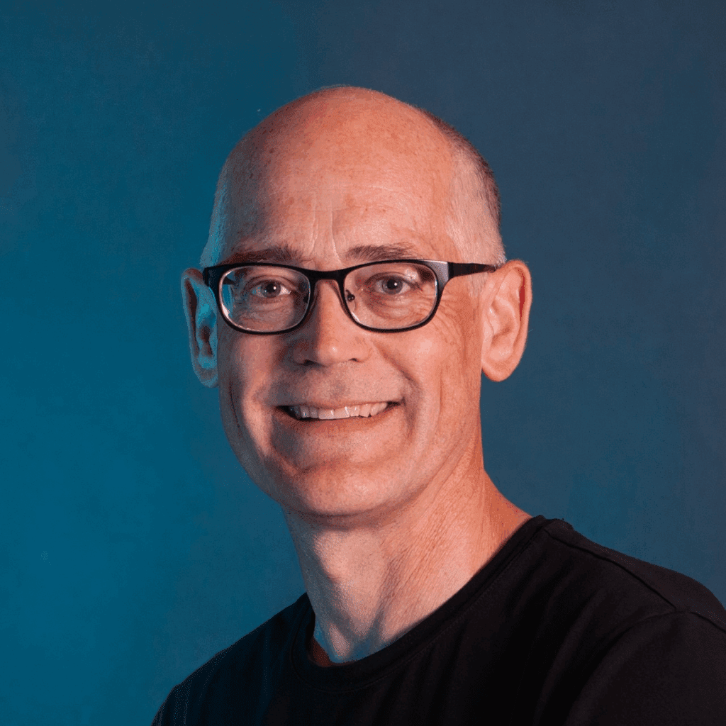 A middle-aged man with glasses and a friendly smile wears a black Trackunit Next shirt, posing against a plain blue background, related to telematics.