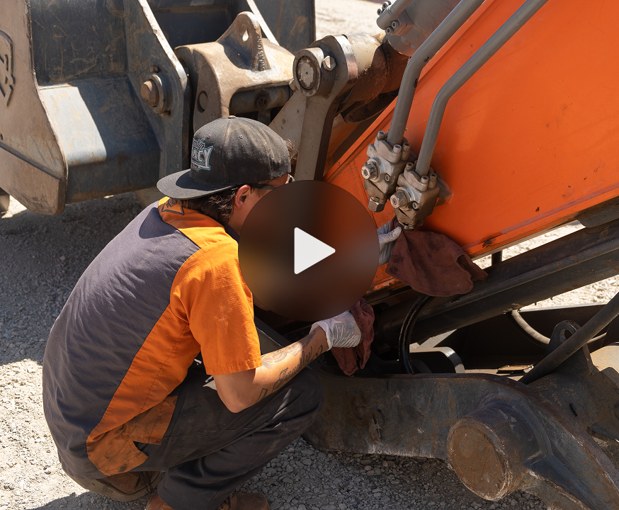 A worker in orange and navy uniform kneels on gravel to service hydraulic lines of an orange construction vehicle, showing fleet management.