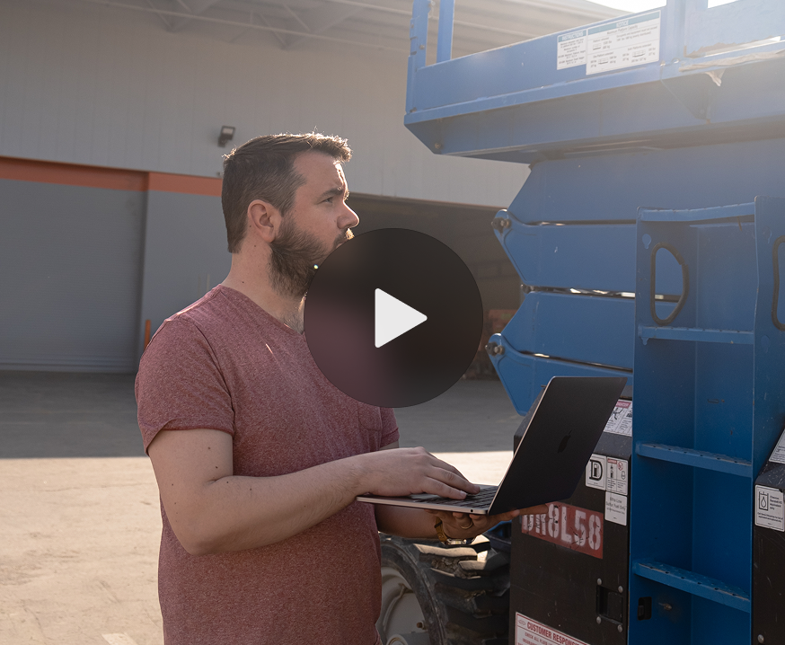 A bearded man in a red t-shirt uses a laptop by blue industrial equipment outdoors, suggesting construction equipment tracking.