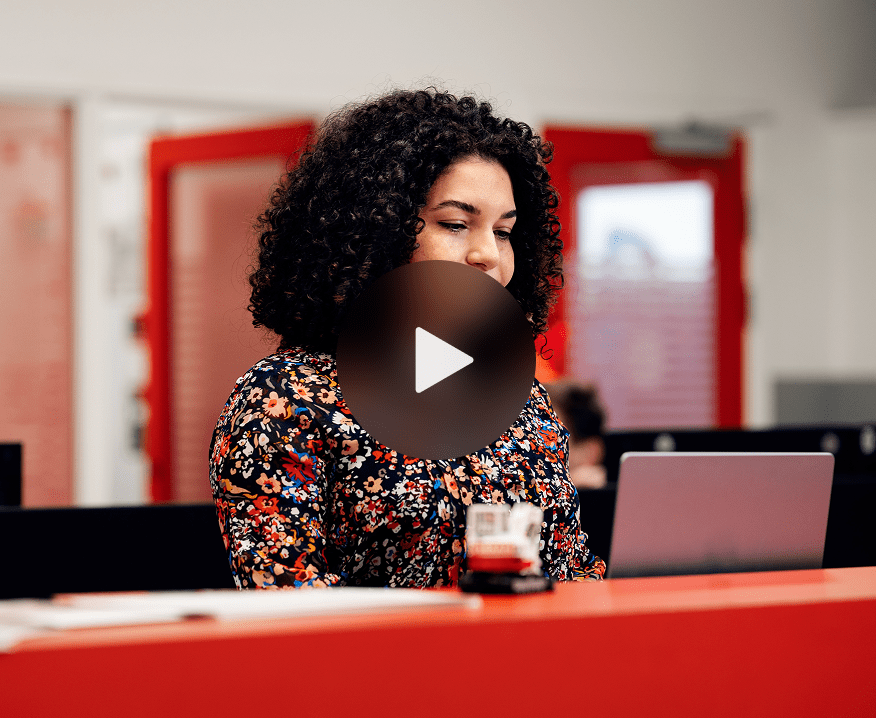 A person with curly hair and a floral top works on a laptop at a red desk in a modern, tech-driven connected jobsite office.