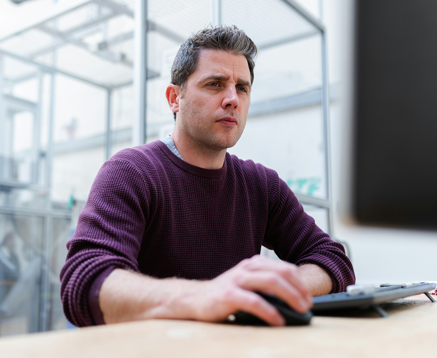 A man in a maroon sweater works at a desk, focused on his monitor in a modern workspace, suggesting connected jobsite activity.