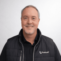 A middle-aged man with short gray hair and a beard smiles, wearing a black polo and Trackunit vest, against a light background.