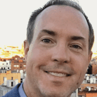 A man with short brown hair smiles at the camera outdoors, buildings and a sunset sky behind him, on a connected jobsite.