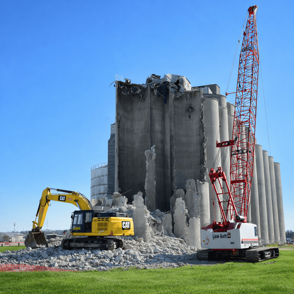 A yellow excavator and red crane demolish concrete silos amid rubble, under a clear blue sky, showing a connected jobsite.