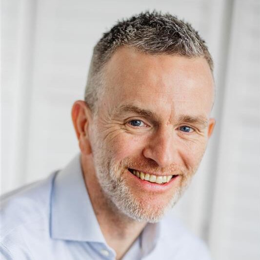 A man with short gray hair and a beard smiles at the camera in a light blue shirt, representing construction equipment tracking.