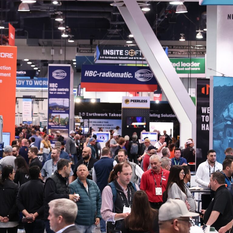 A large crowd at a busy trade show explores colorful booths and banners, networking and learning about construction IoT solutions.