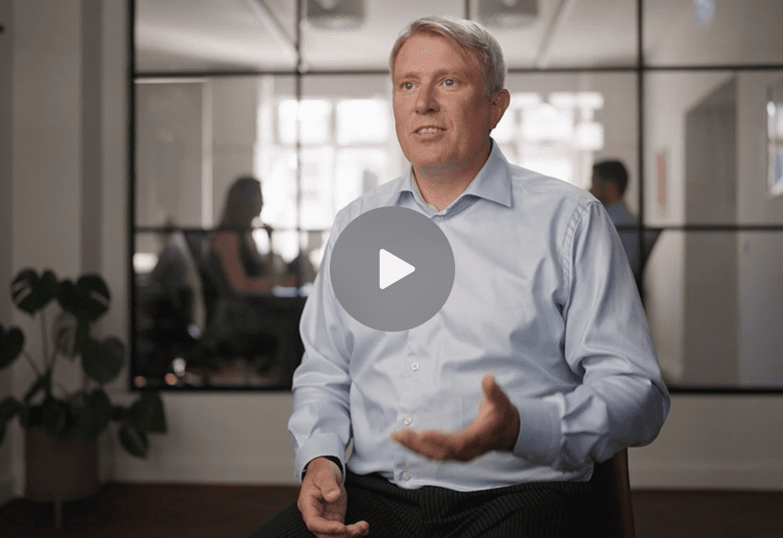 A man in a light blue shirt gestures while speaking in an office, with coworkers and a play button overlay, highlighting connected jobsite.