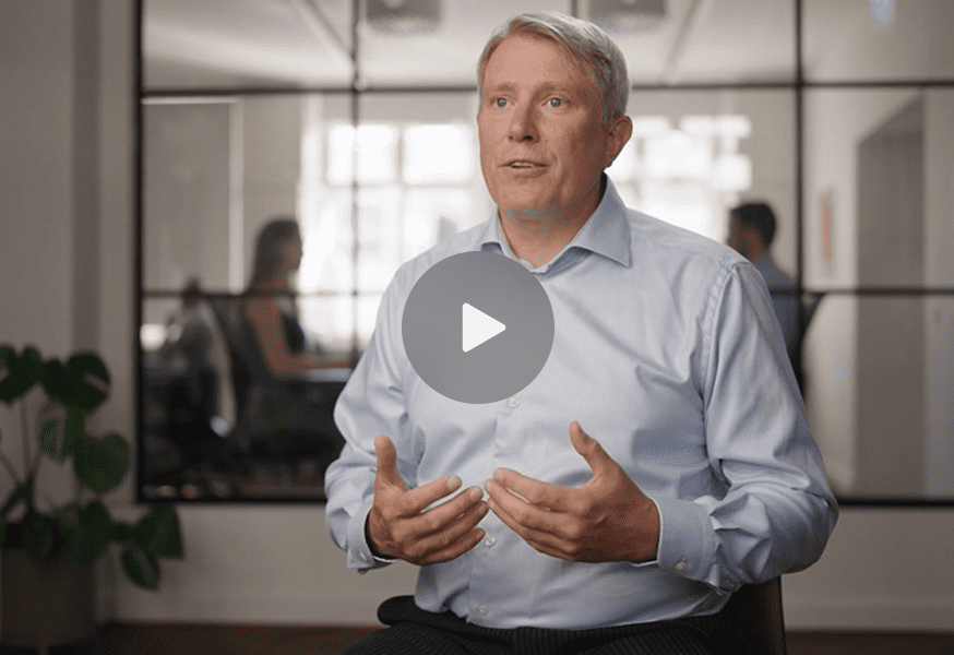 A man in a light blue shirt speaks and gestures in an office, with two people working behind a glass wall, suggesting a connected jobsite.