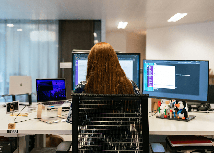 A person with long red hair works at a desk with several monitors and a laptop in a modern office, focusing on construction IoT tasks.