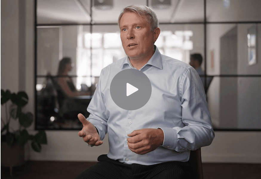 A man in a light blue shirt gestures while speaking in an office, with a play button overlay; blurred figures sit behind a glass wall, suggesting fleet management.