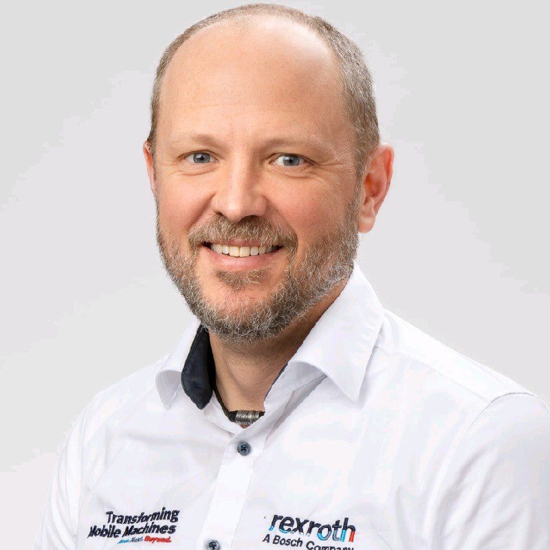 A smiling man with short hair and a beard in a collared shirt with company logos poses against a plain light background.